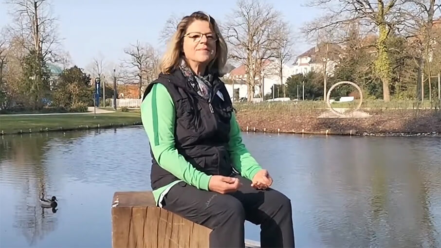 Frau sitzt auf vor einem Teich und zeigt Atemübung